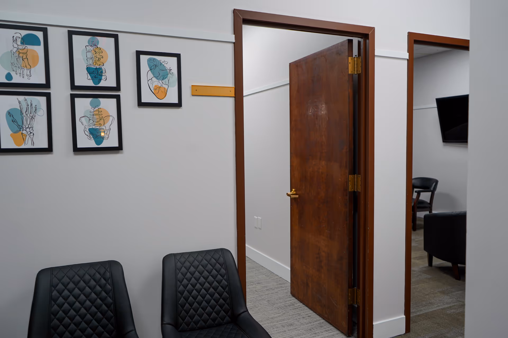 Hallway in a senior facility with framed art, open door, and black upholstered chairs.
