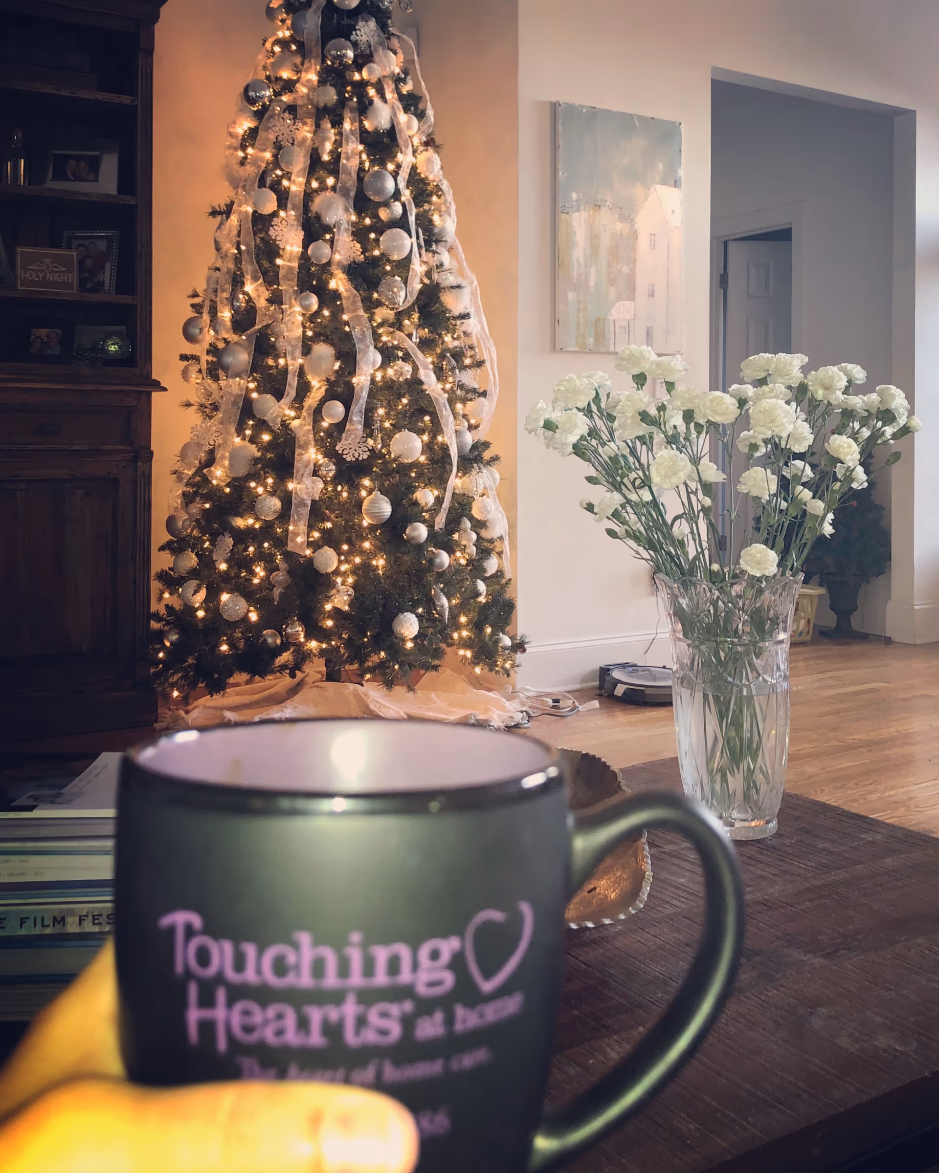A Christmas tree with ornaments and a vase of white flowers in a cozy living room.