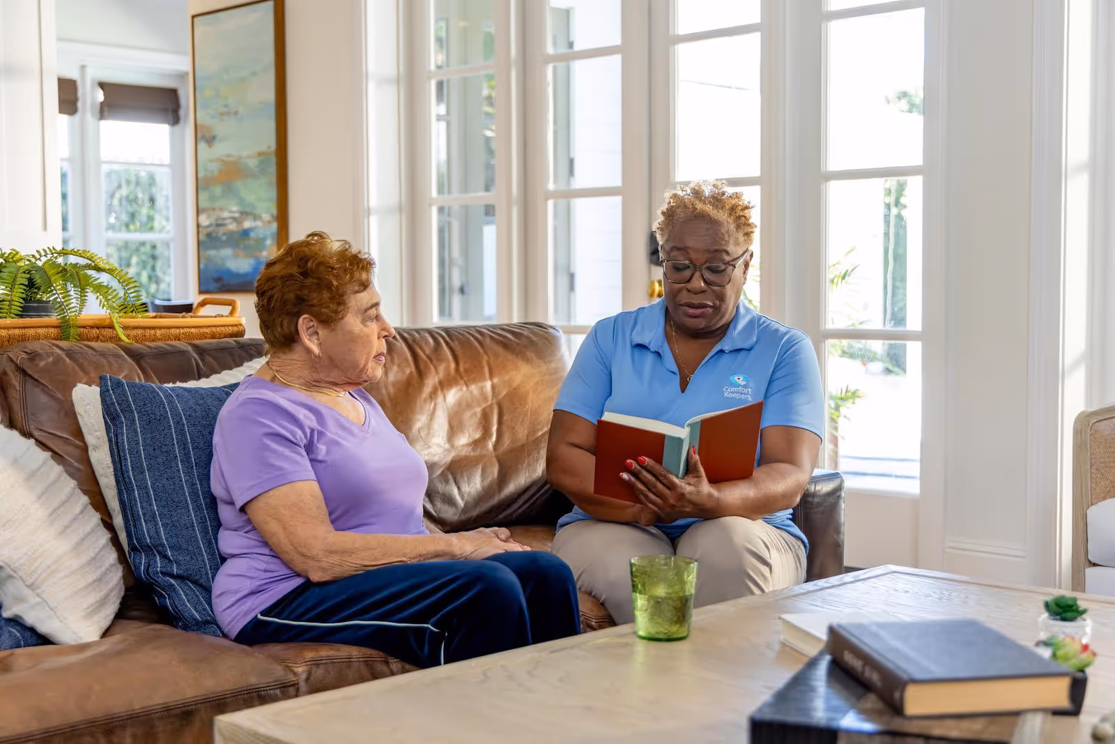 Caregiver reads aloud to elderly woman while seated on a brown leather living room sofa.