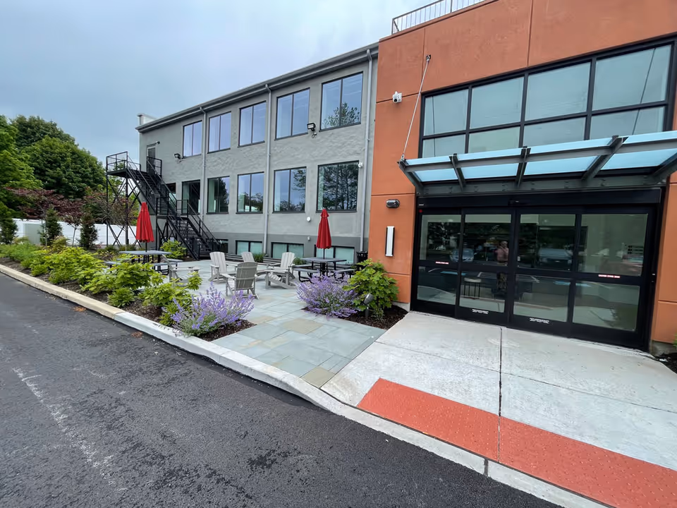 Outside patio with tables, chairs, red umbrellas, and landscaped plants beside the building.
