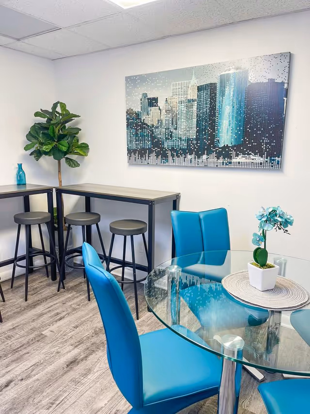 Bright dining area with glass round table, blue chairs, potted orchid, and cityscape art.