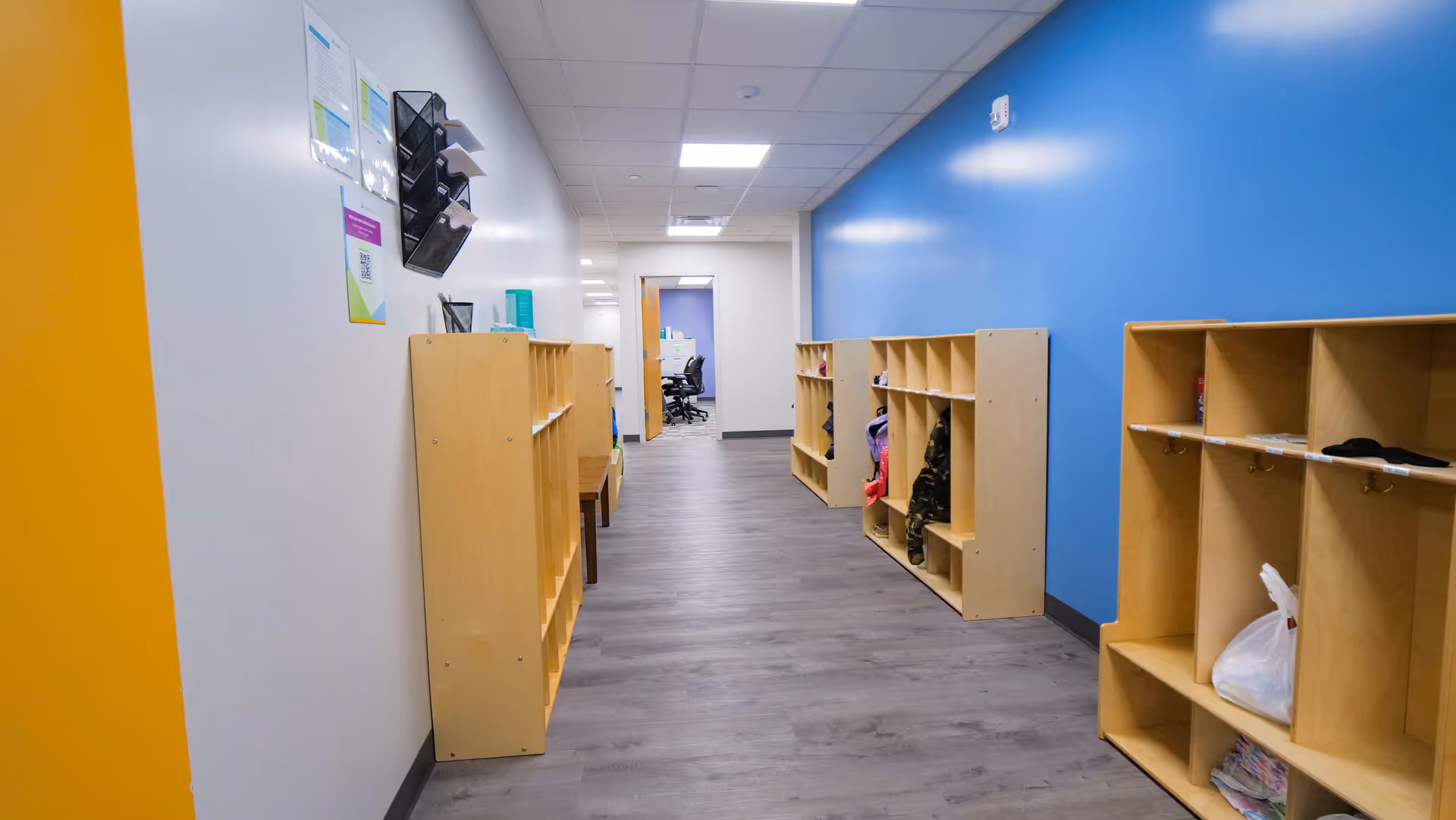 A bright hallway featuring wooden cubbies along a blue wall and grey flooring.