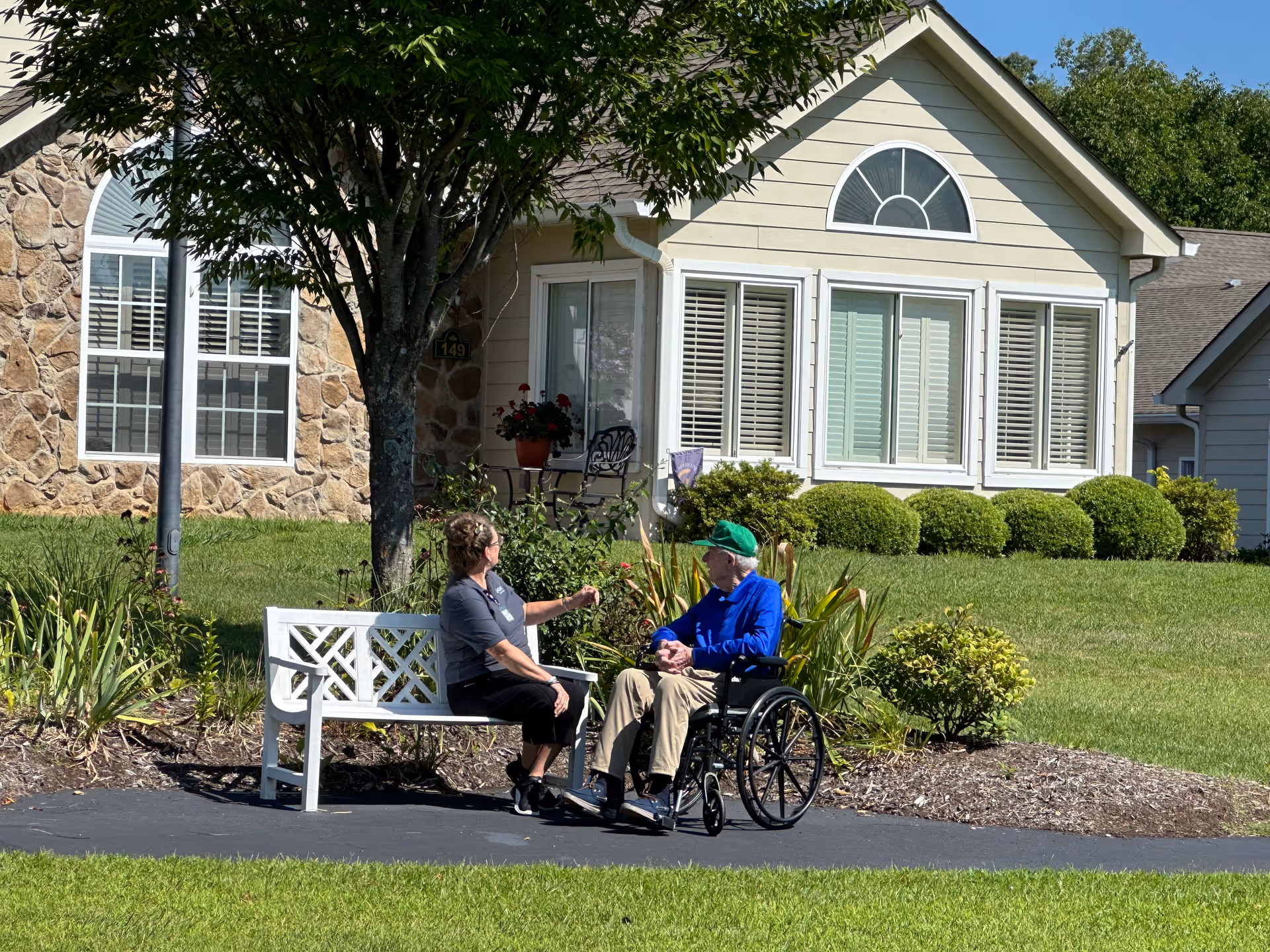 Two people sit on a bench beside a wheelchair outside a beige residential facility with landscaping.