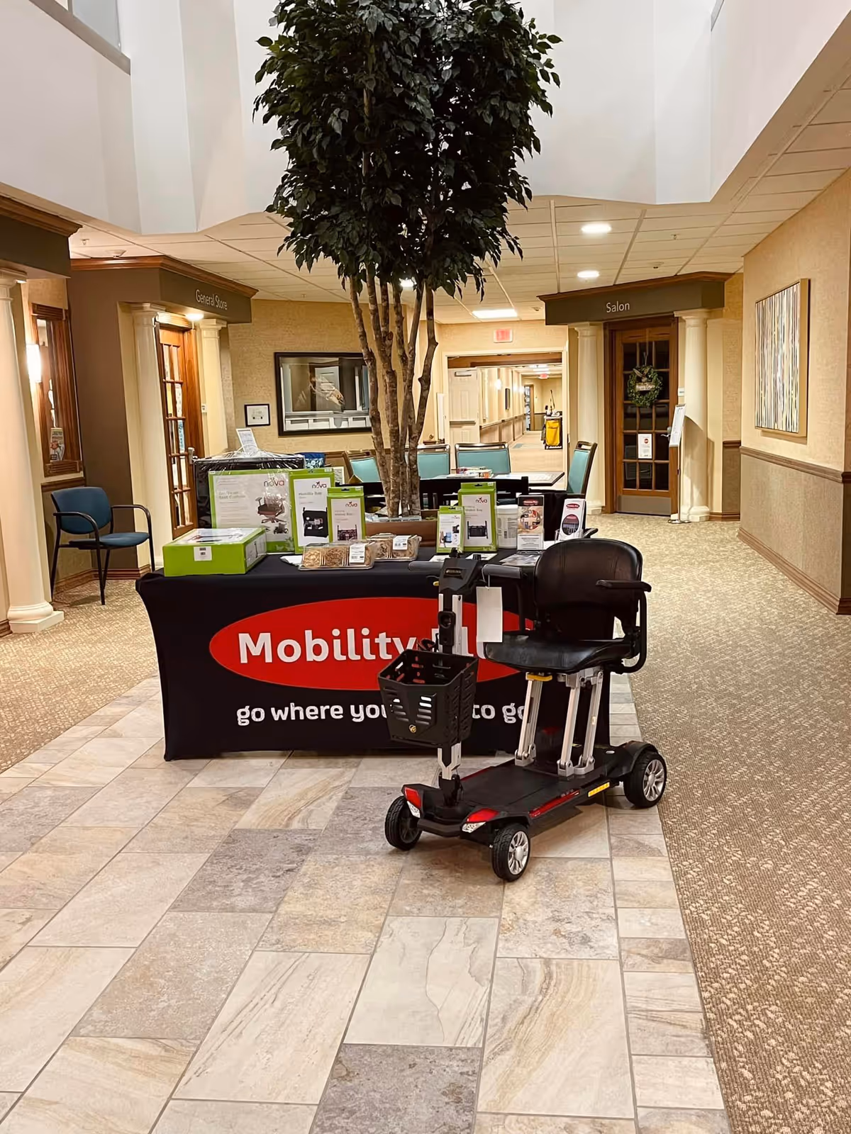 Indoor senior living lobby with Mobility Plus display table and a mobility scooter