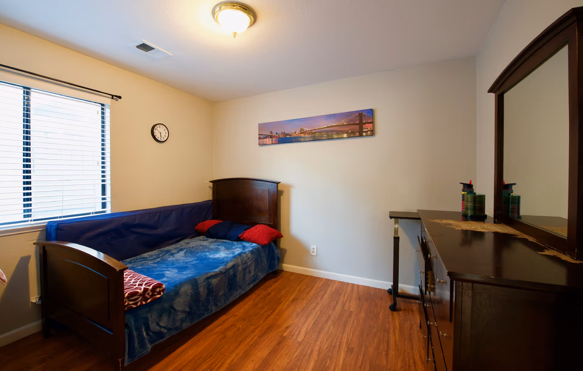 A bedroom with a dark wood bed, blue bedding, dresser, window blinds, and wall clock.