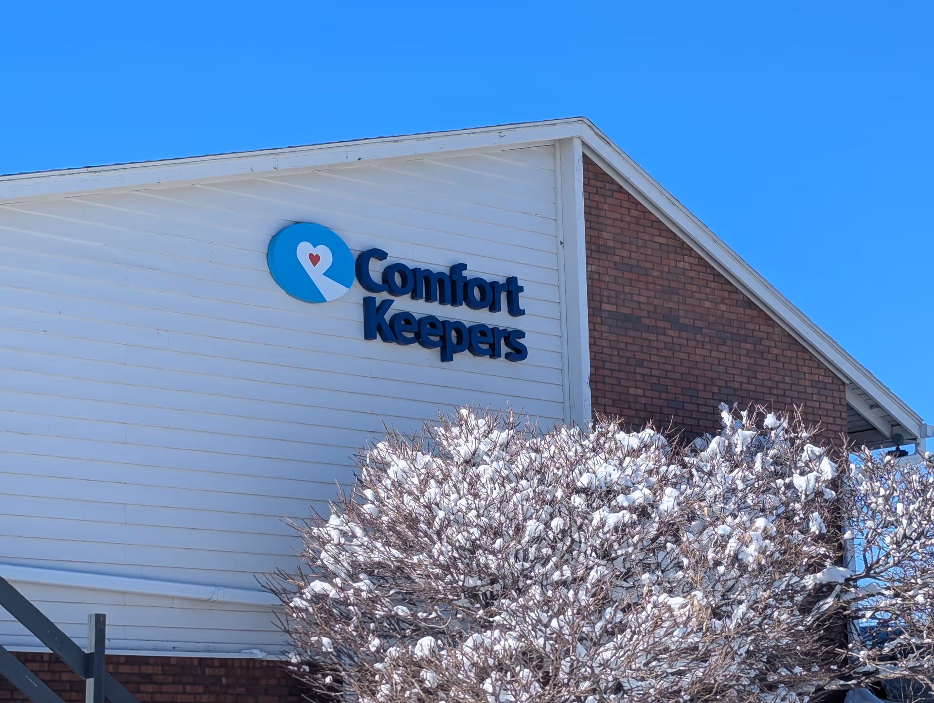 Exterior view of a Comfort Keepers building against a blue sky with snow on bushes.