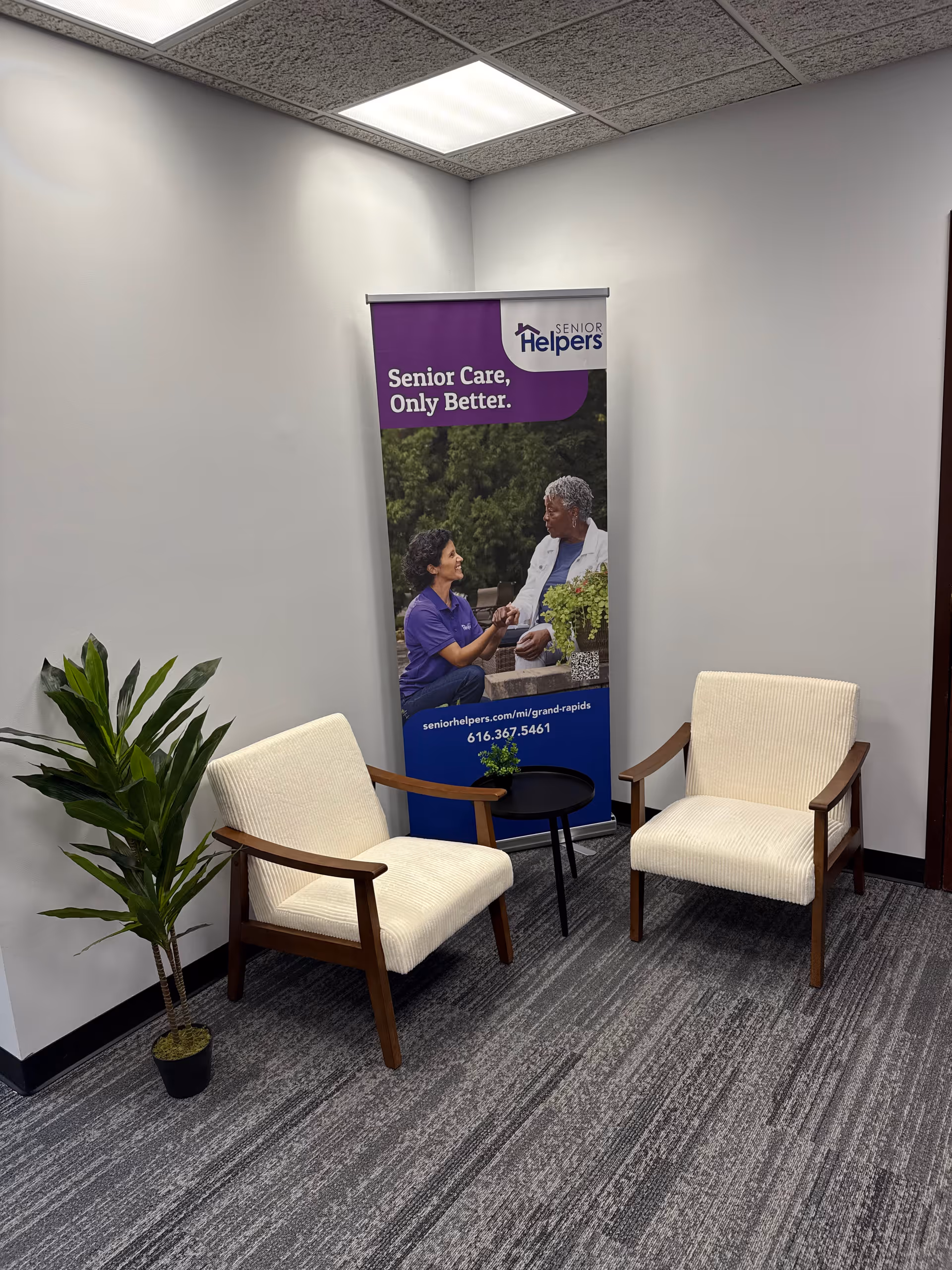 A small waiting area with two cream chairs, a plant, and a Senior Helpers banner.