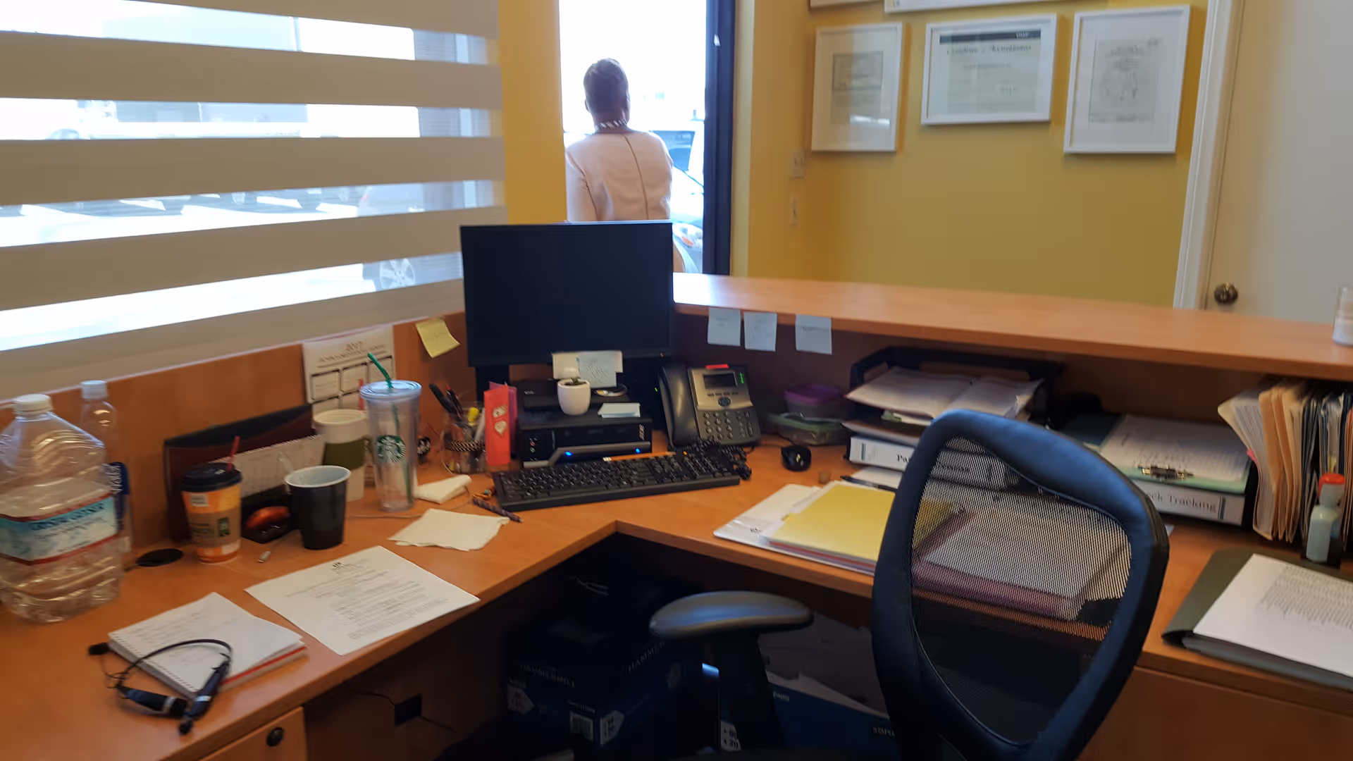 Interior front desk with computer, scattered papers, drinks, and a person near the doorway.