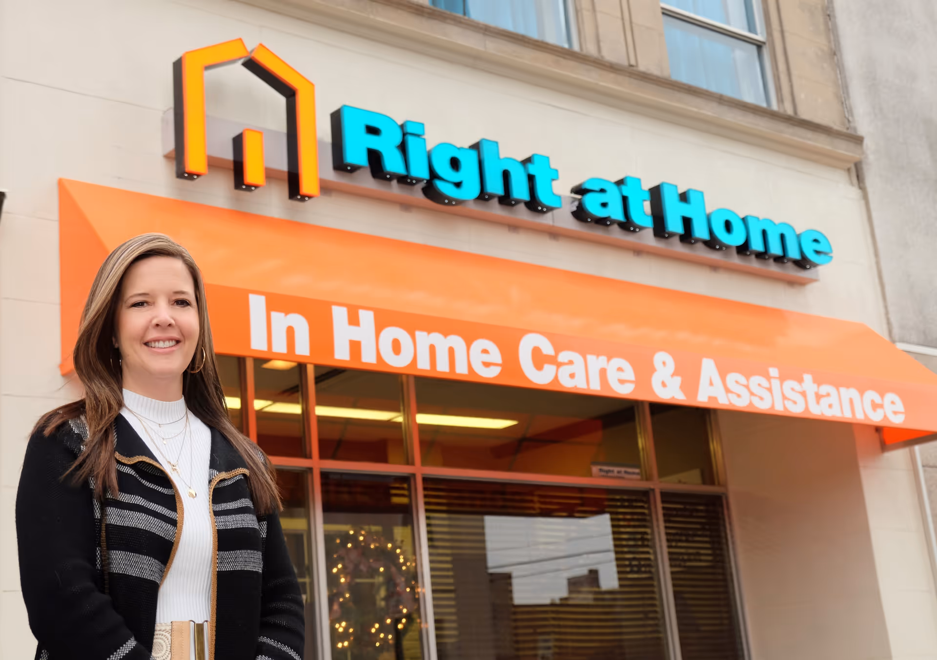A smiling woman stands in front of a Right at Home storefront with an orange awning.