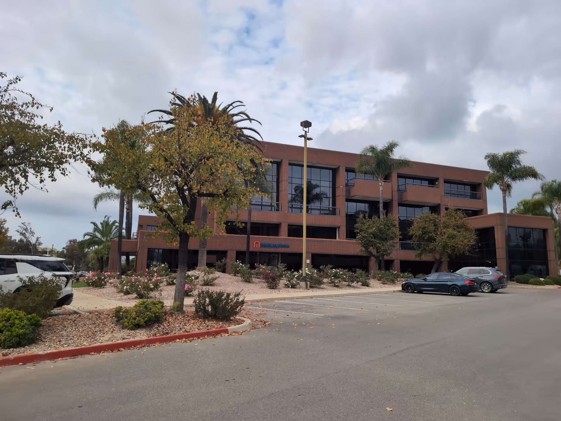 Brick senior living building with glass windows, palm trees, and a parking lot in front.