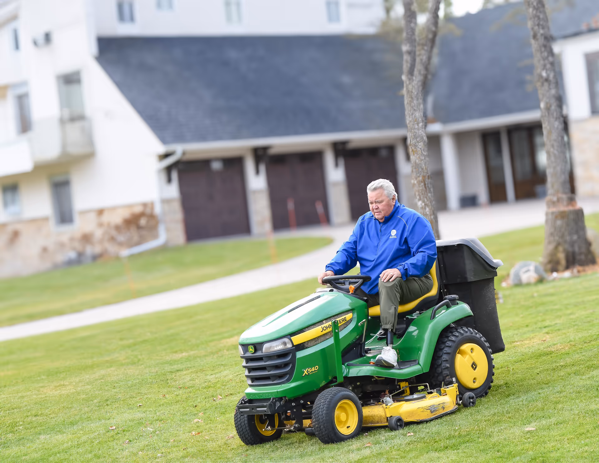 An elderly man operates a green lawn mower on a grassy area beside a large building.