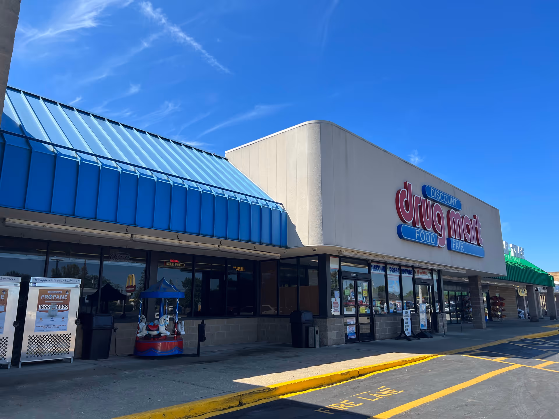 Exterior view of a Discount Drug Mart storefront with blue awning and parking lot.