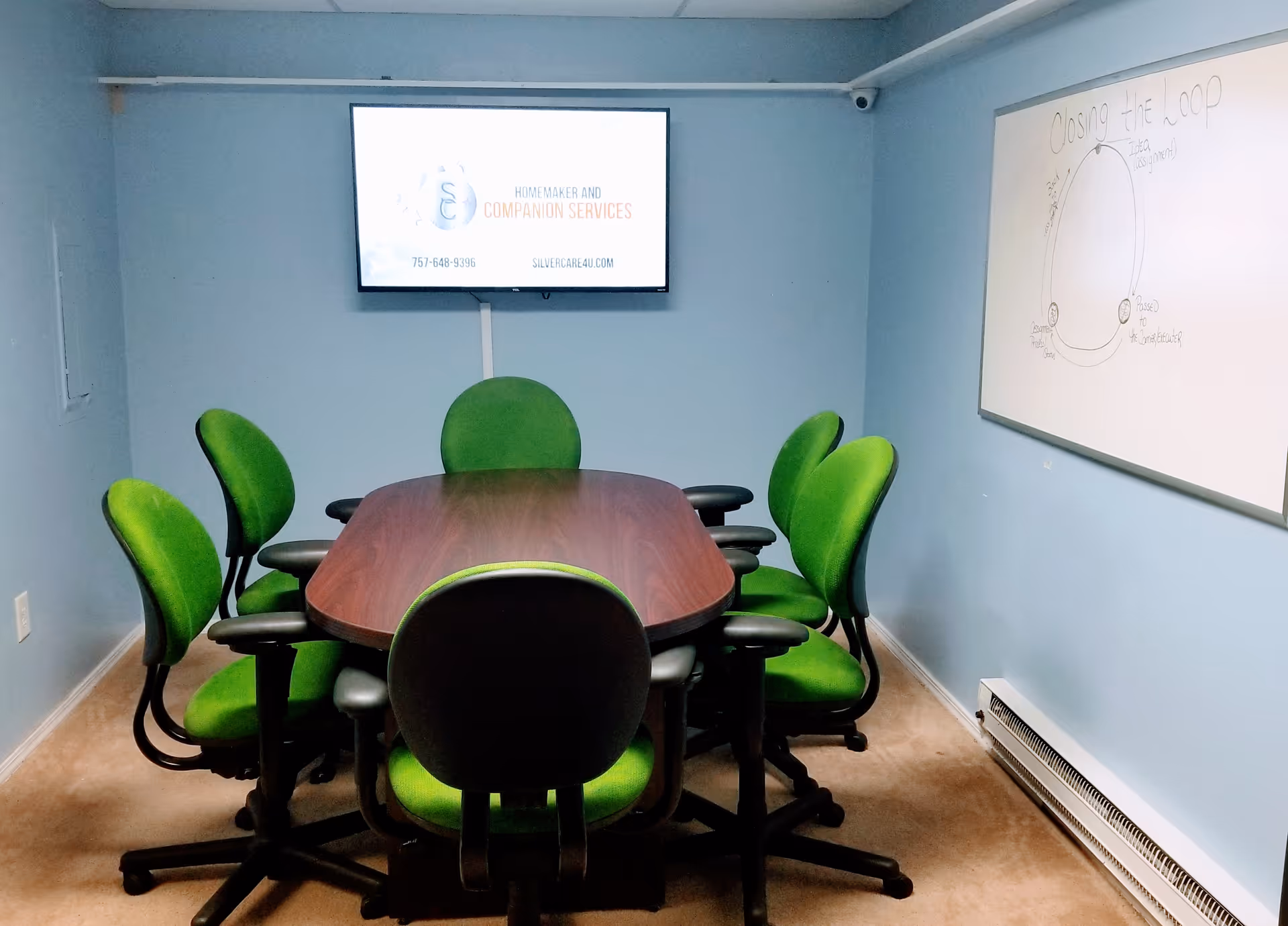 Small conference room with a wooden table and bright green chairs, whiteboard, and wall-mounted screen.