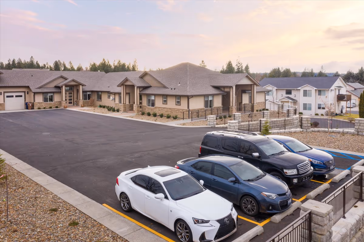Exterior view of a senior living campus with beige buildings, parked cars, and a paved lot.