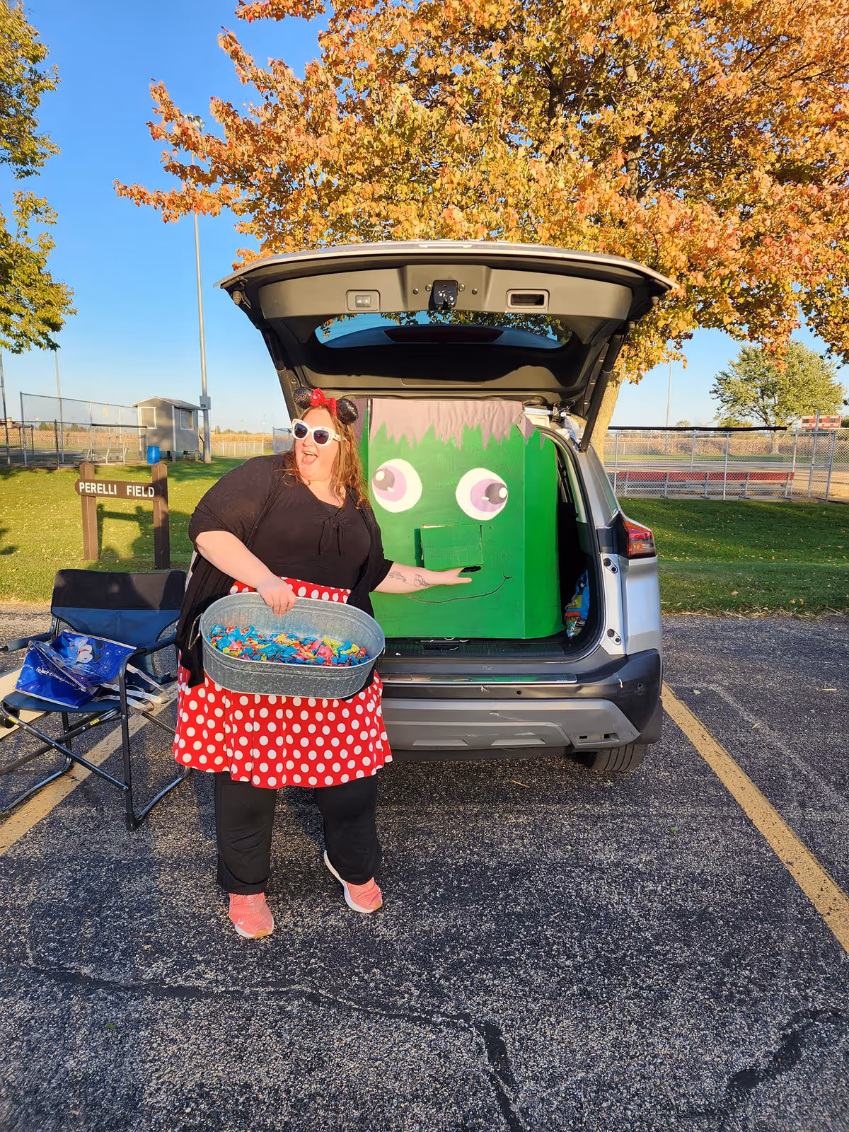Woman in red polka-dot skirt stands beside an open SUV trunk outdoors, holding candy.