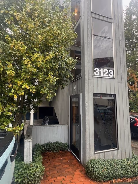 Exterior entrance of a grey two-story building with glass doors, 3123, and greenery.