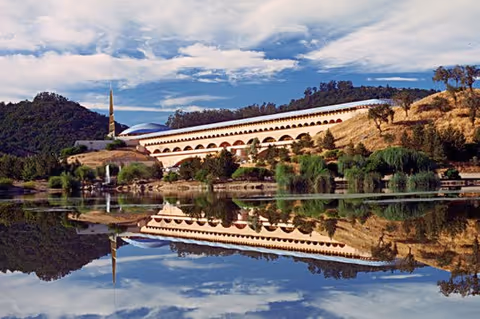 Serene waterfront view of an arched beige building on a hillside, reflected in calm water.