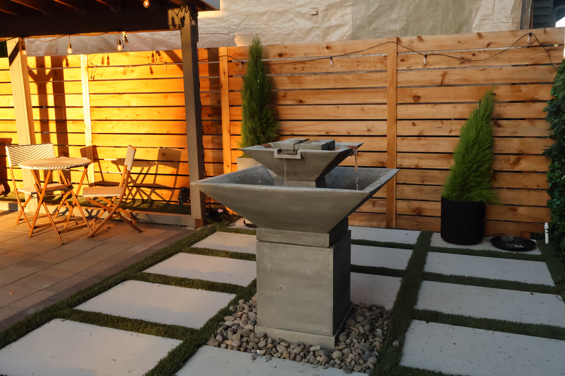 Outdoor courtyard with a modern water fountain, wooden fence, string lights, and seating.