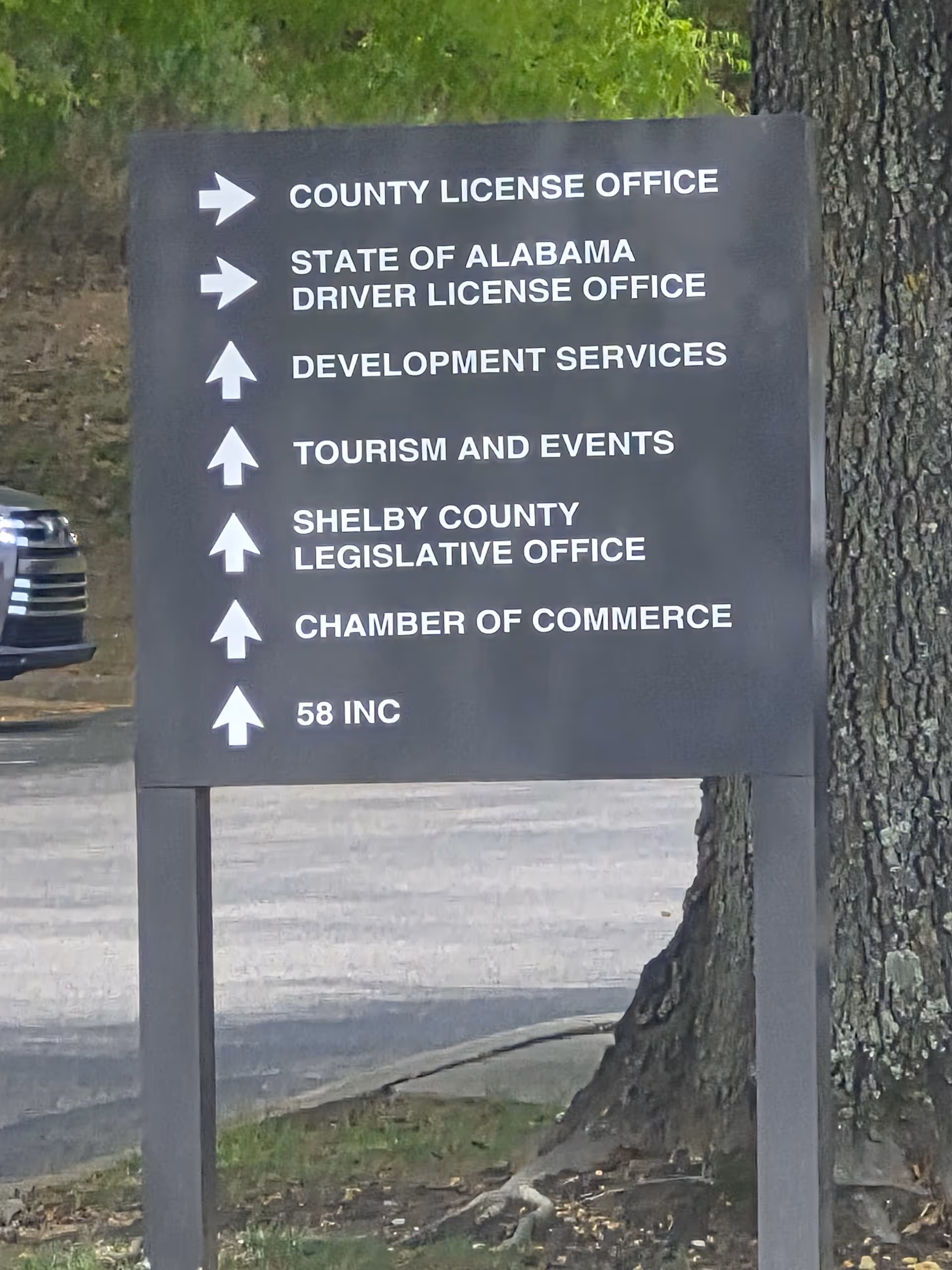 Outdoor sign listing county offices with arrows beside a tree in a parking lot.
