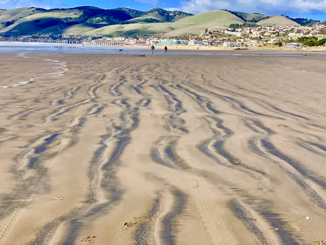Wide beach with ripple-sand patterns, ocean, and distant hills along the shore.
