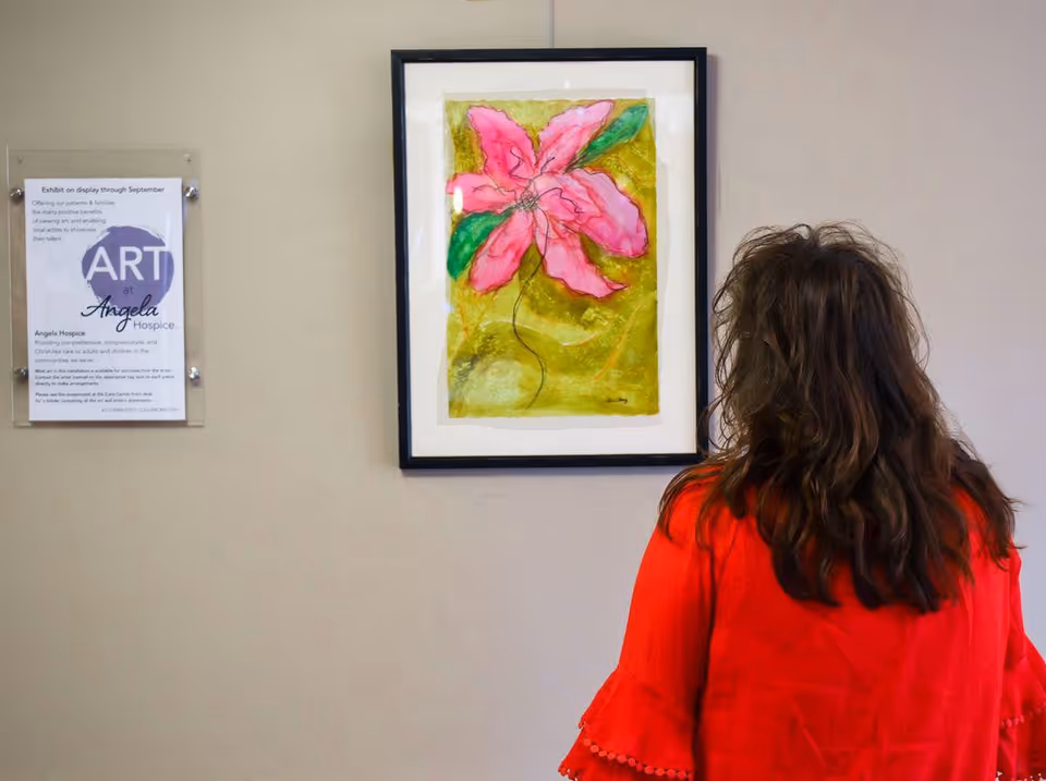 A person in red stands with back to camera, admiring a framed pink flower painting.