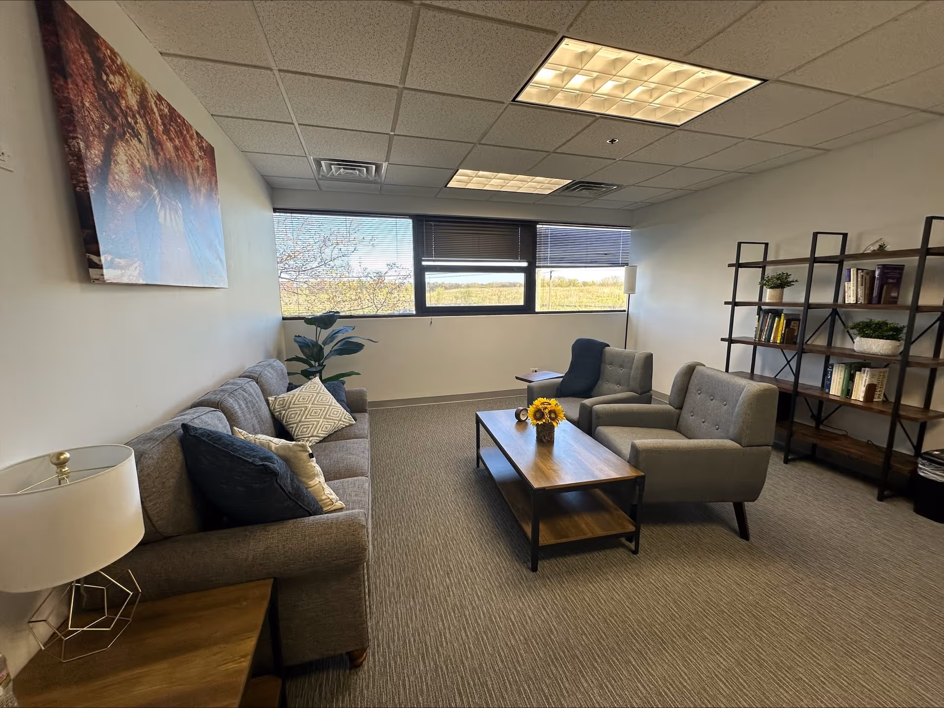 Modern senior living lounge with gray couches, chairs, coffee table, bookshelf, and window view.