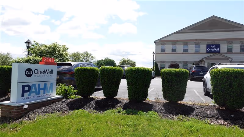 Exterior view of OneWell Health Care facility with signage, shrubs, and parked cars.