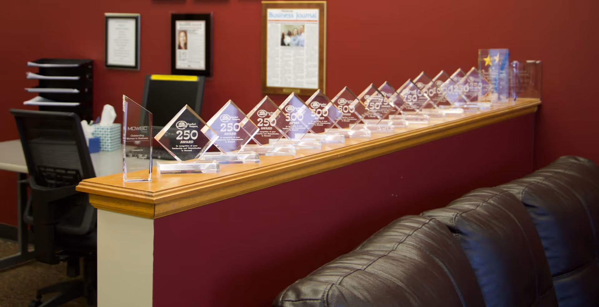 Reception counter lined with glass award plaques against a burgundy wall and brown leather sofa.