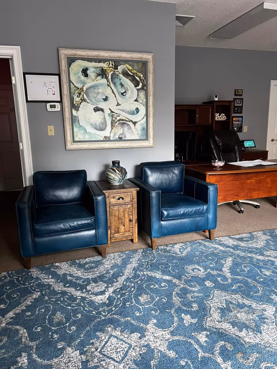 Two blue leather chairs flank a wooden side table beside a painting in a reception area.