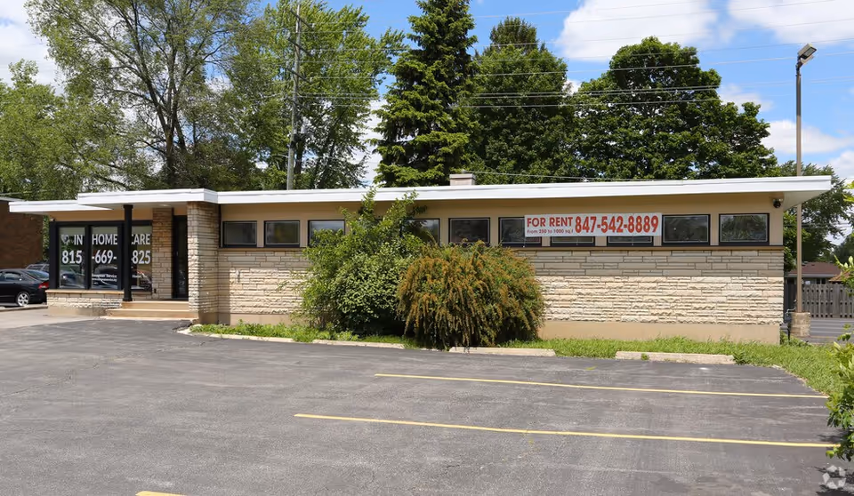Single-story brick building front with large windows, 'For Rent' sign, and parking lot.