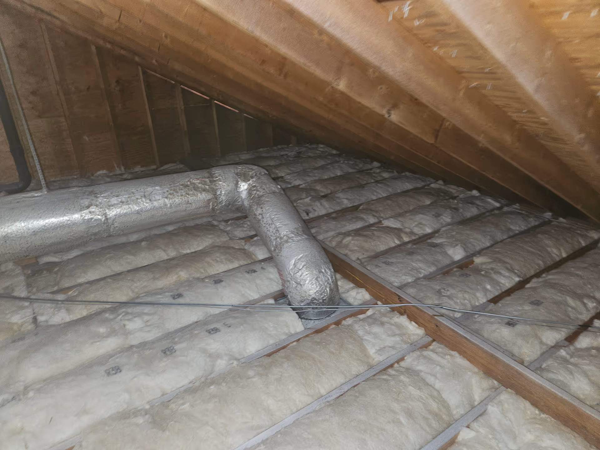 Attic space showing exposed rafters, insulation, and ductwork between wooden beams.