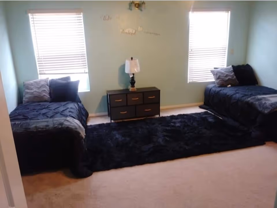 Two twin beds with dark bedding flank a dresser and lamp in a light blue bedroom.