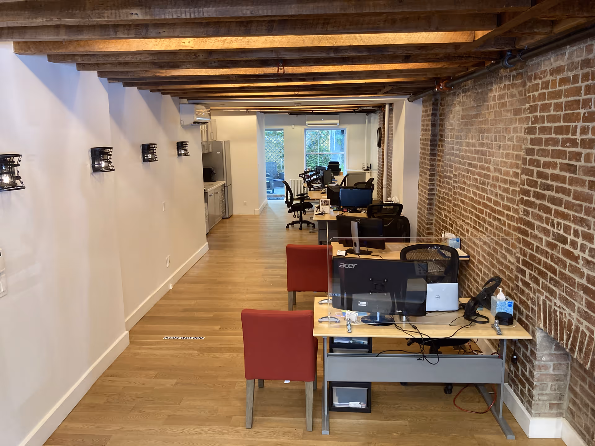 Long interior office area with multiple desks, computers, and exposed brick walls.