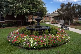 Circular flower bed with a tiered fountain, blooming flowers, and surrounding green lawn.