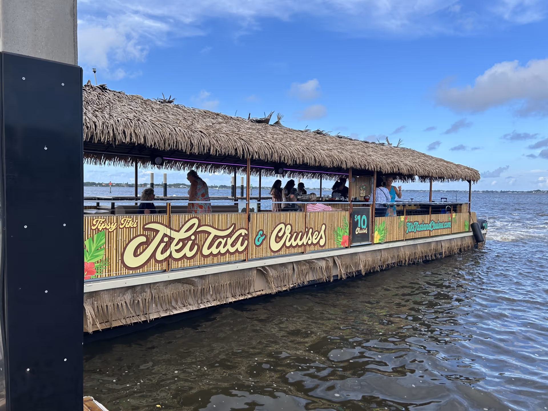 Thatched-roof pontoon boat with passengers cruising on calm water under blue sky.