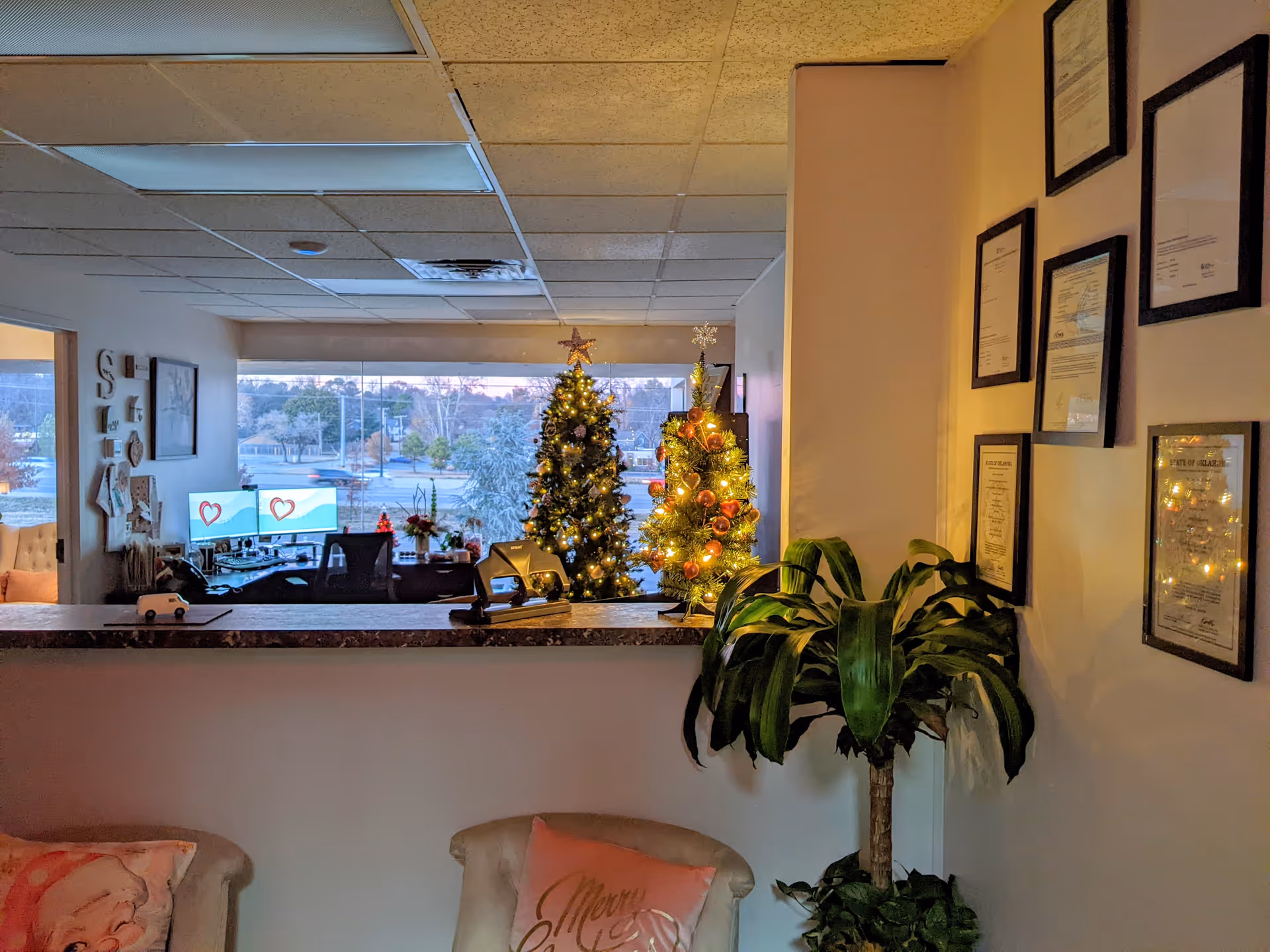 Reception lobby with Christmas trees, computers, a plant, and framed certificates on the wall.