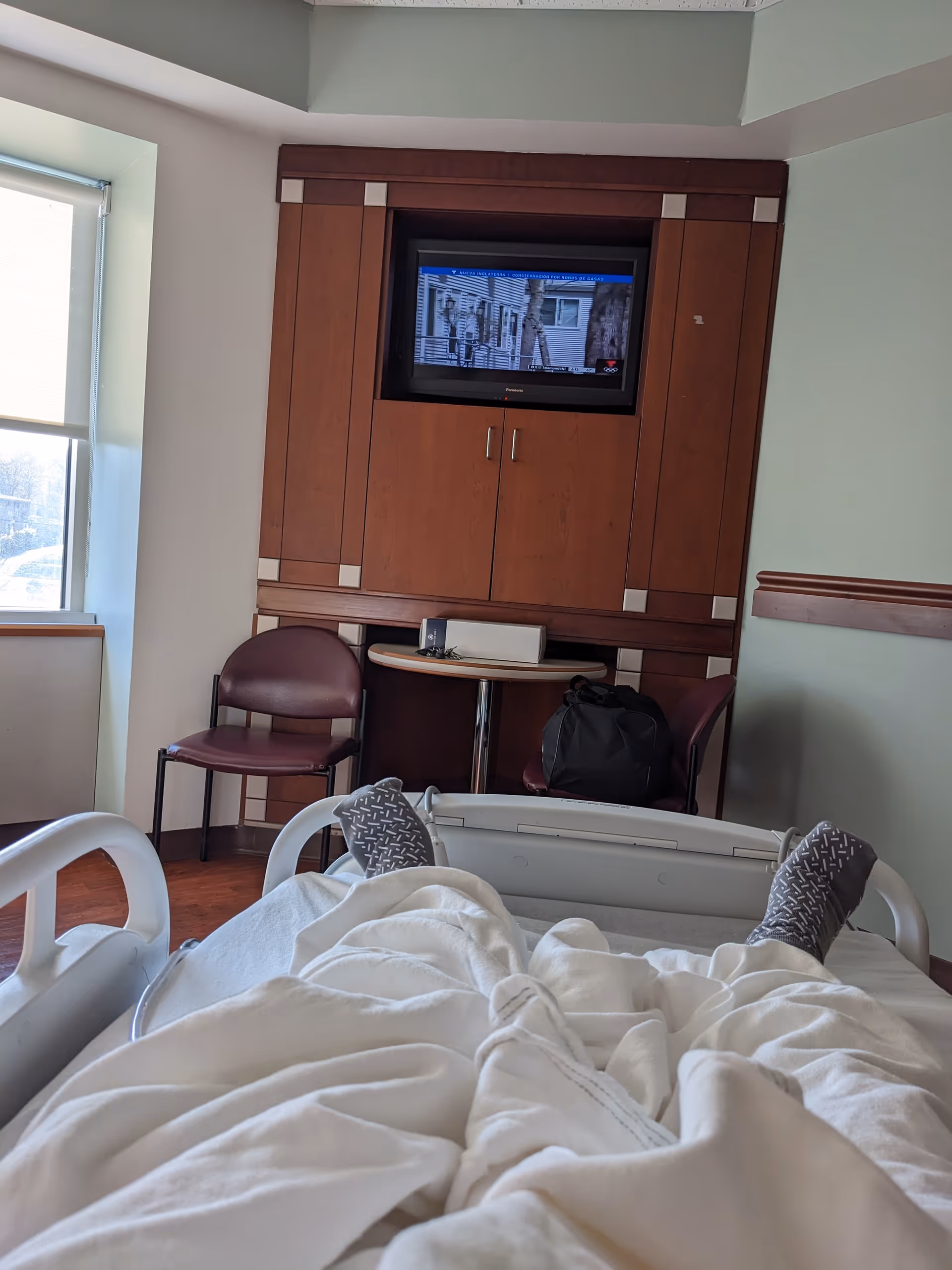 Interior patient bedroom with a bed, blanket, TV in a wooden cabinet, and chairs.