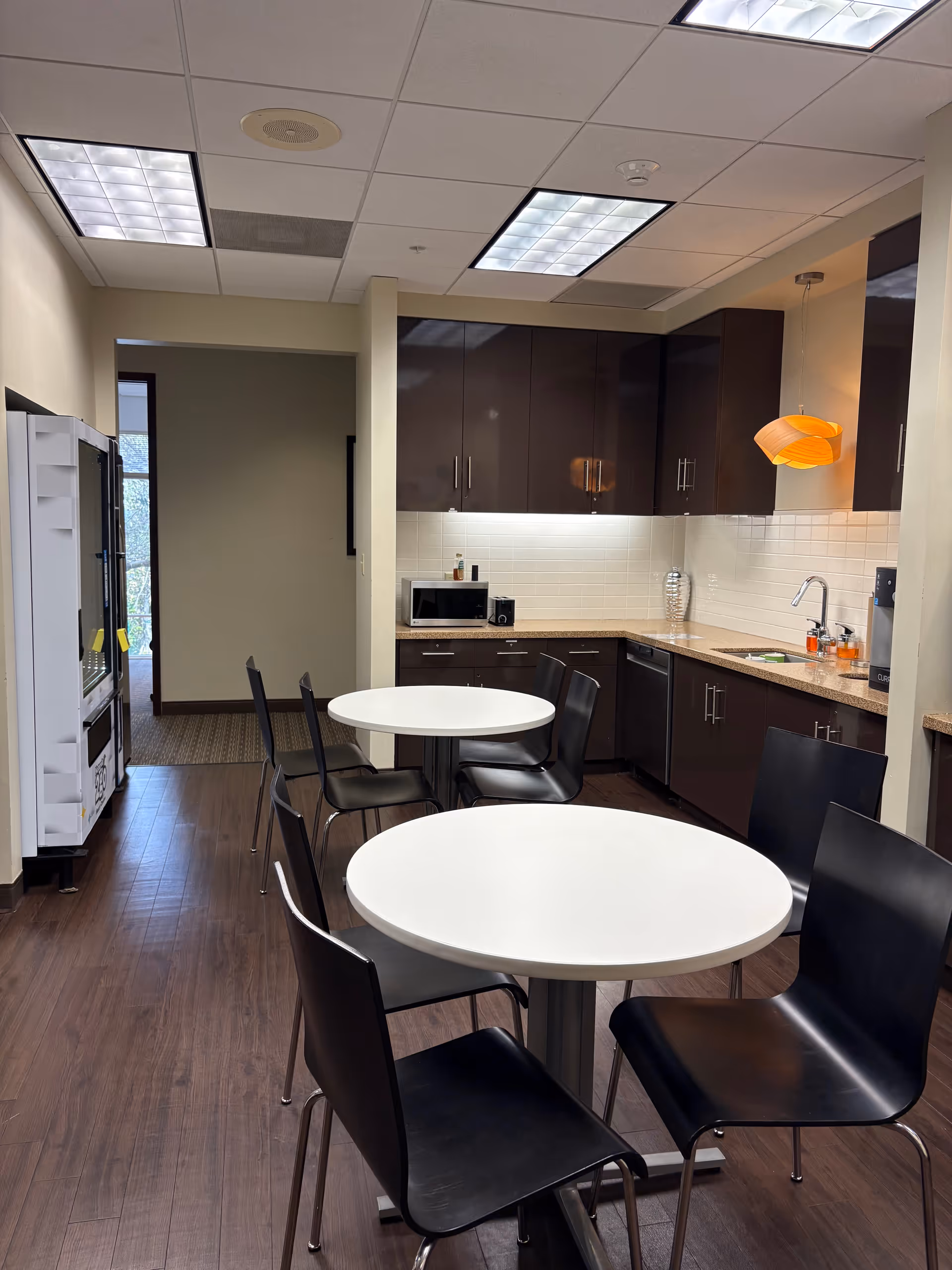 Indoor dining area with round white tables, black chairs, and a kitchenette along the wall.