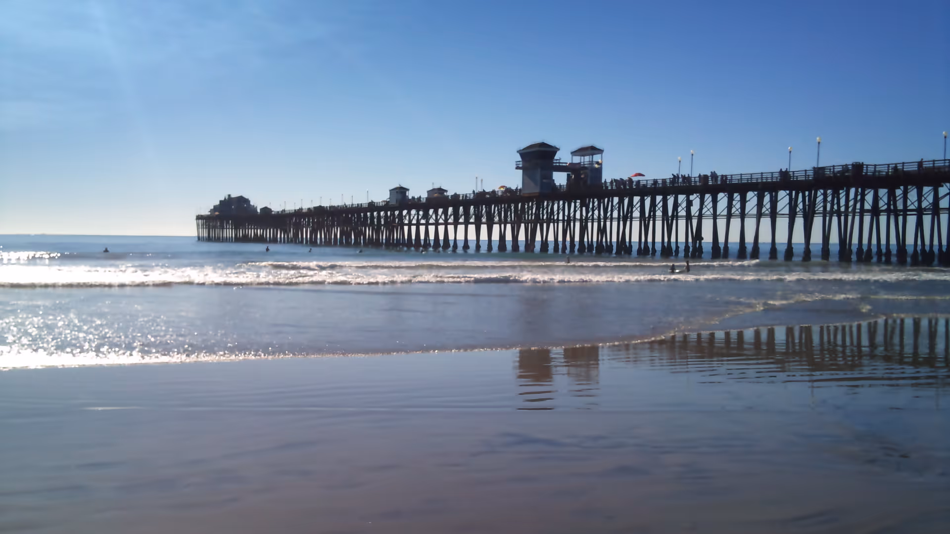 Long wooden pier extends into calm ocean under blue sky, with reflections on wet sand.