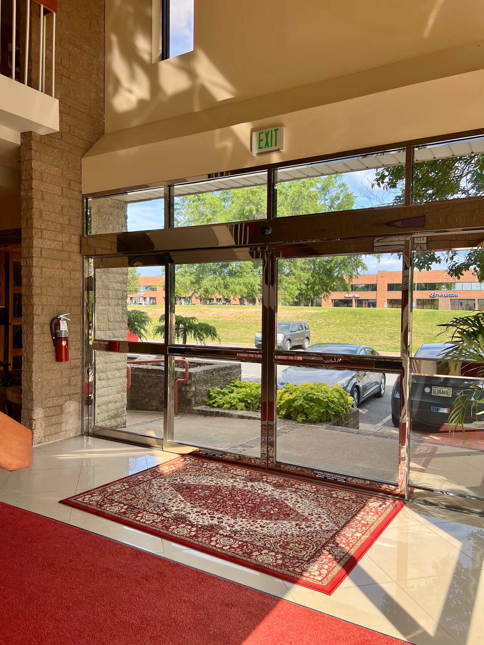 Interior building lobby with glass doors, exit sign, and a red patterned rug.
