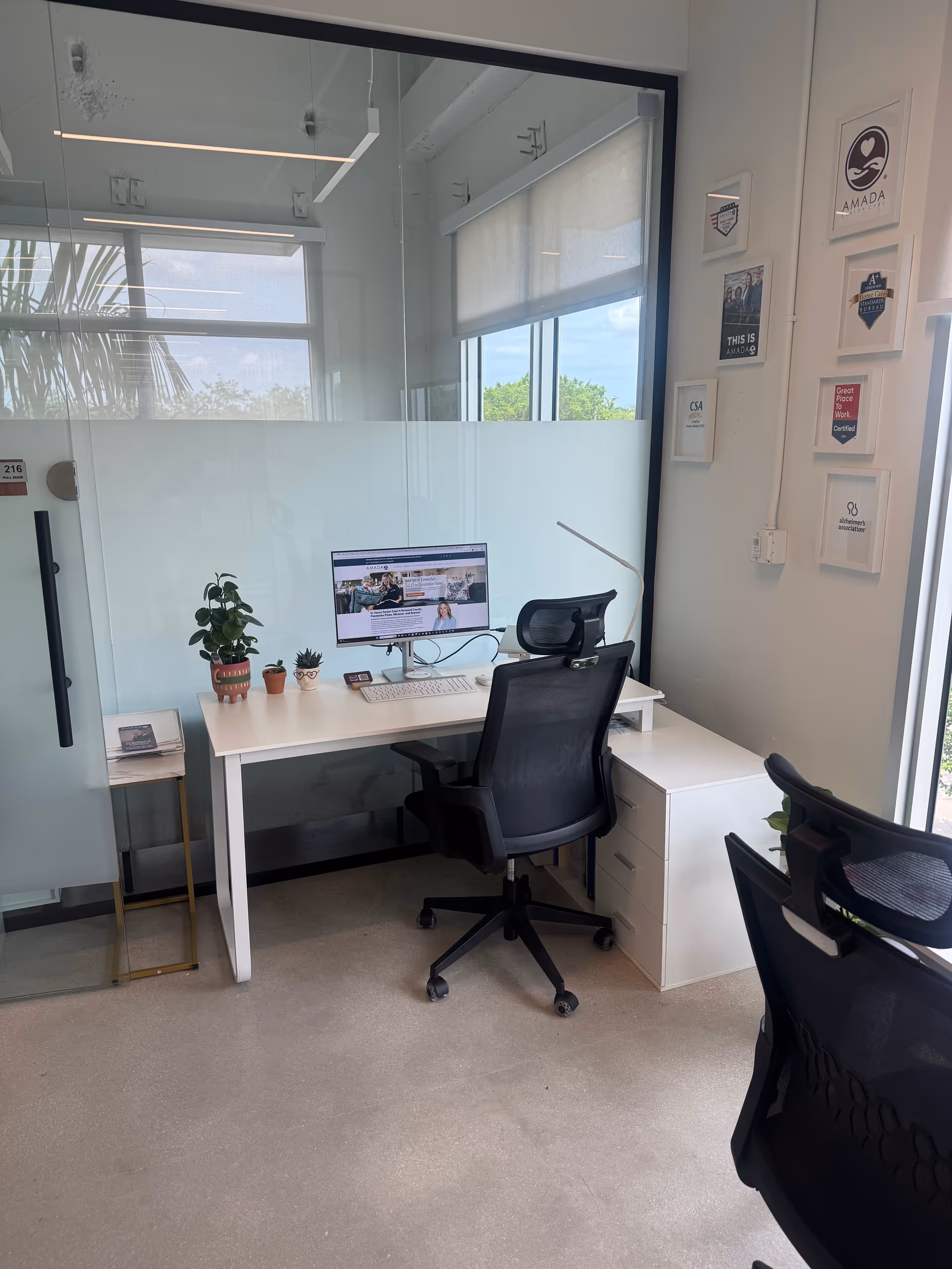 A modern office workspace with a desk, computer, chair, glass walls, and plants.