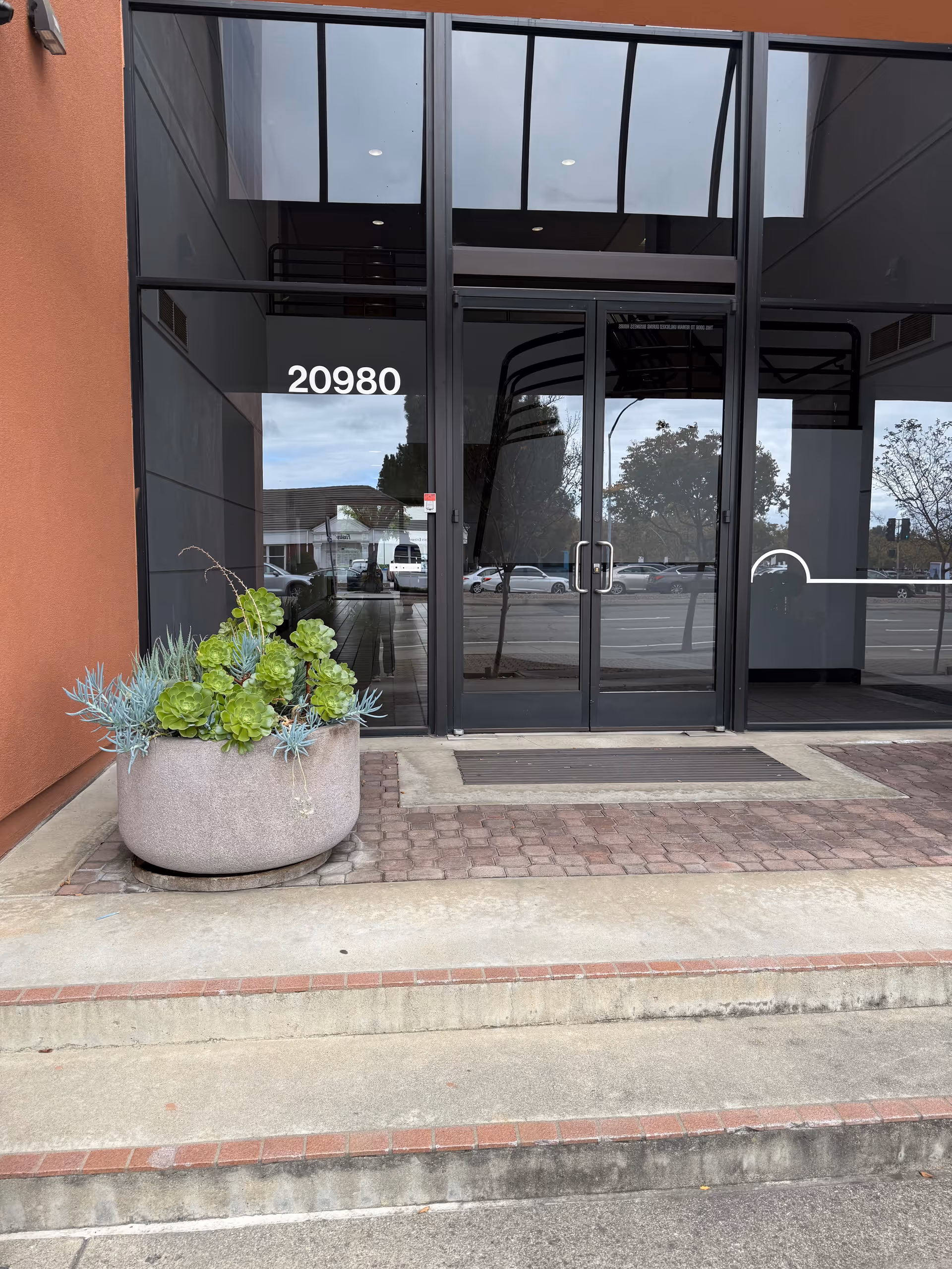 Modern glass entrance with address 20980 and a large planter outside the building front
