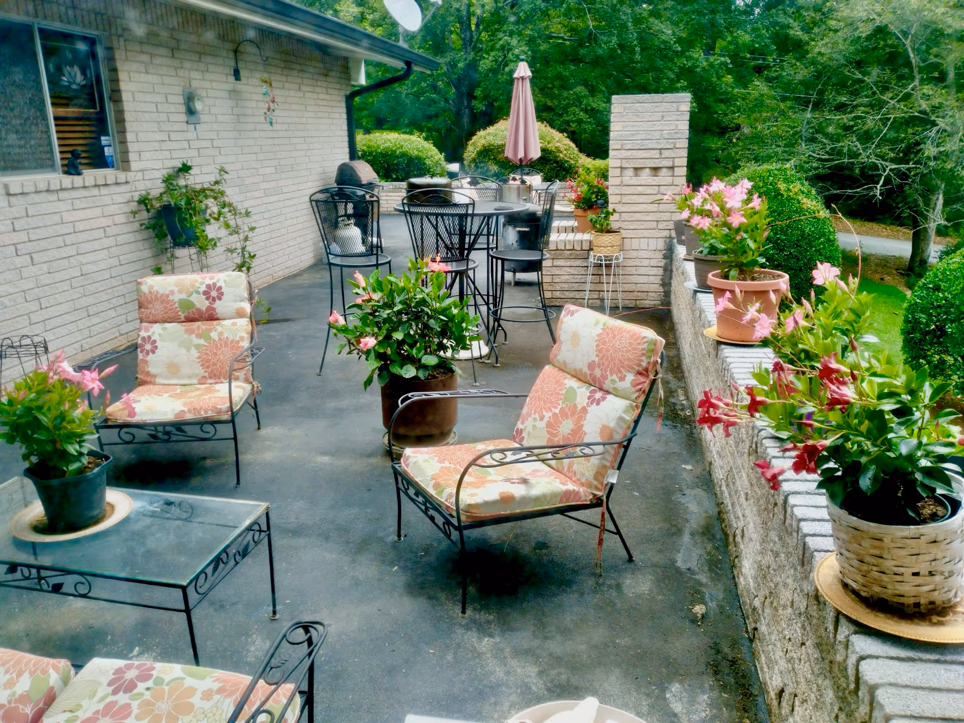 Outdoor patio with cushioned metal chairs, tables, and potted flowers beside a brick wall.