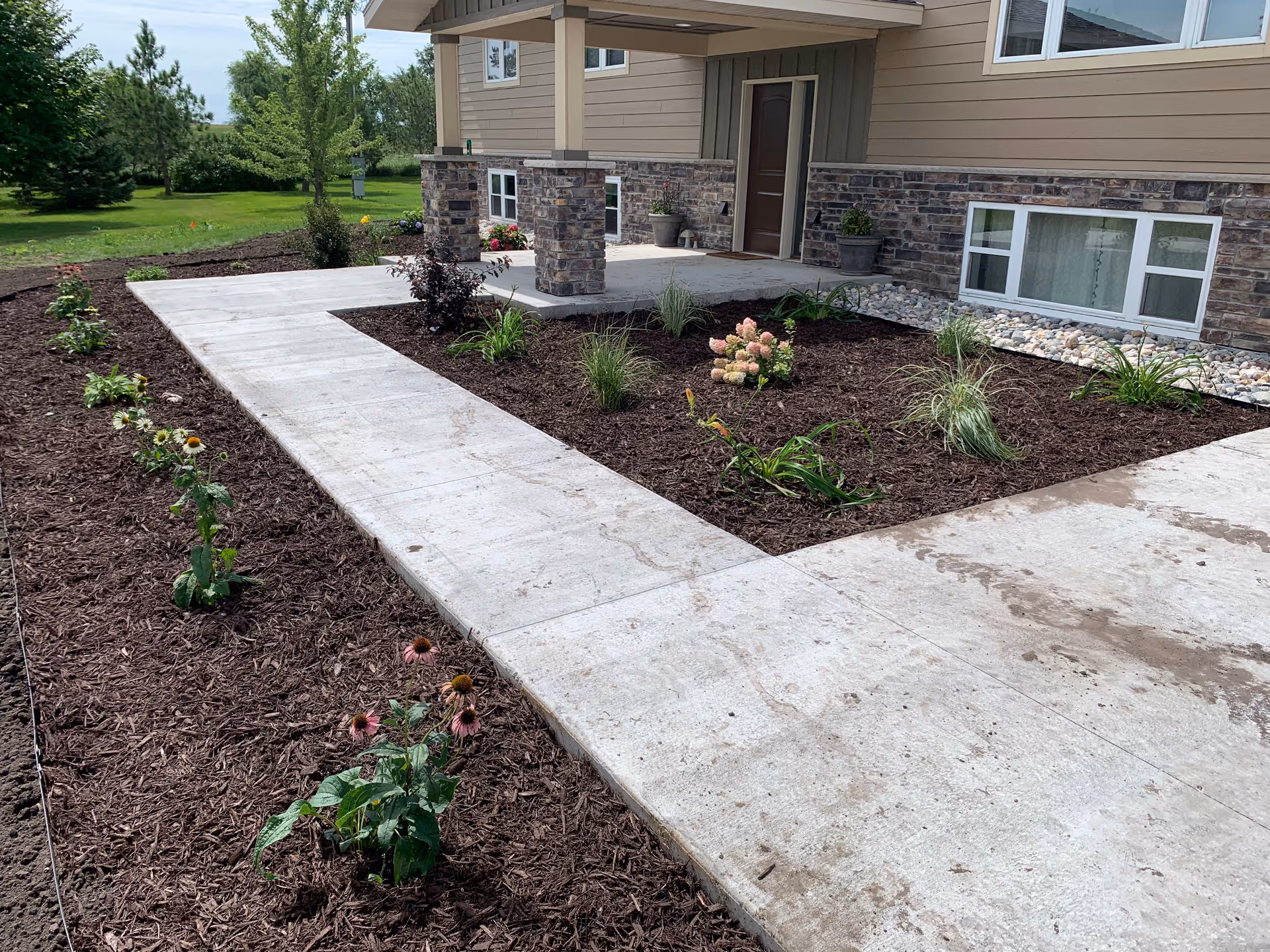 Front entrance of a beige building with stone accents, concrete walkway, and flower beds.