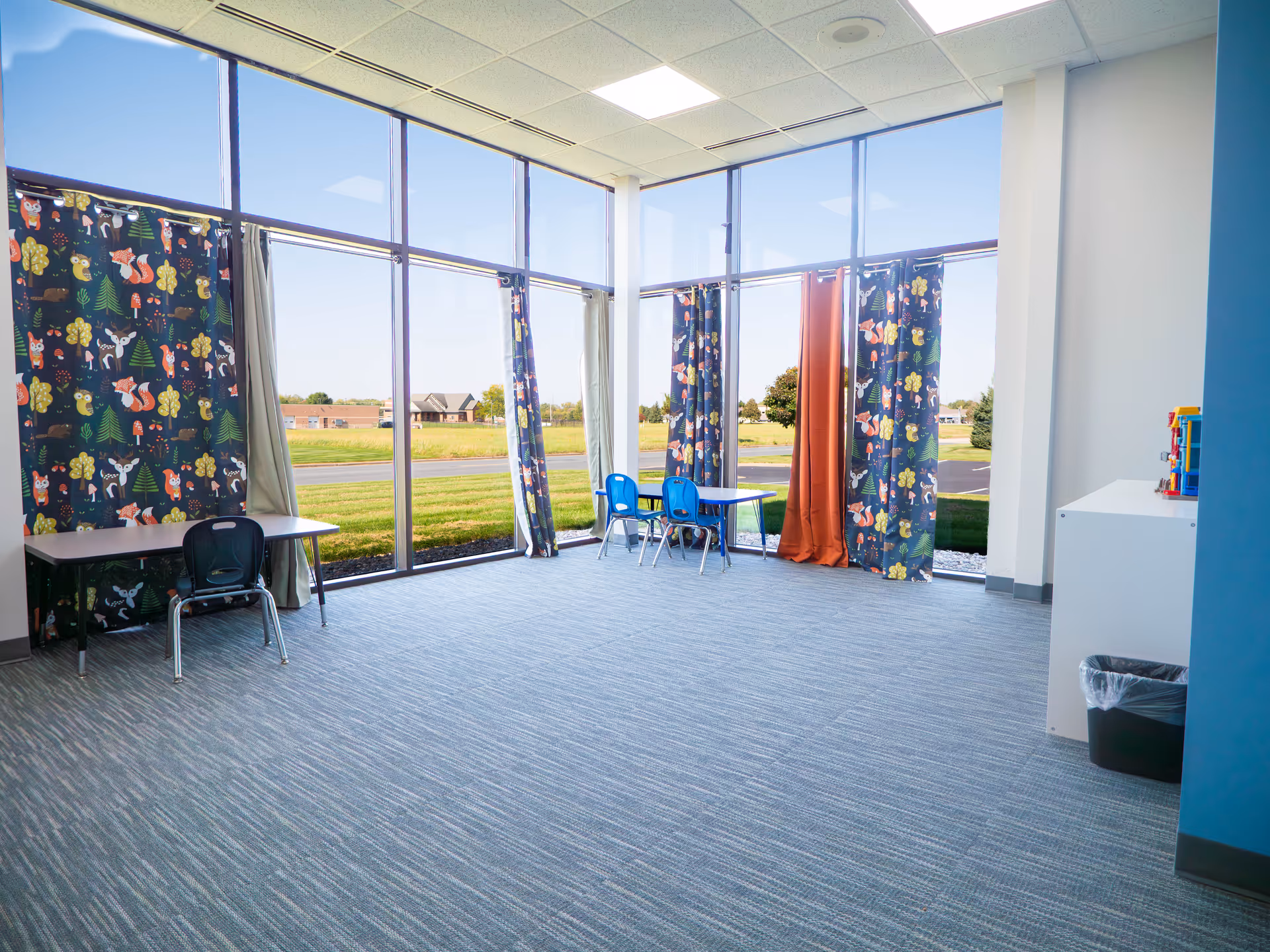 Bright interior play area with large windows, colorful curtains, small tables, and blue chairs.