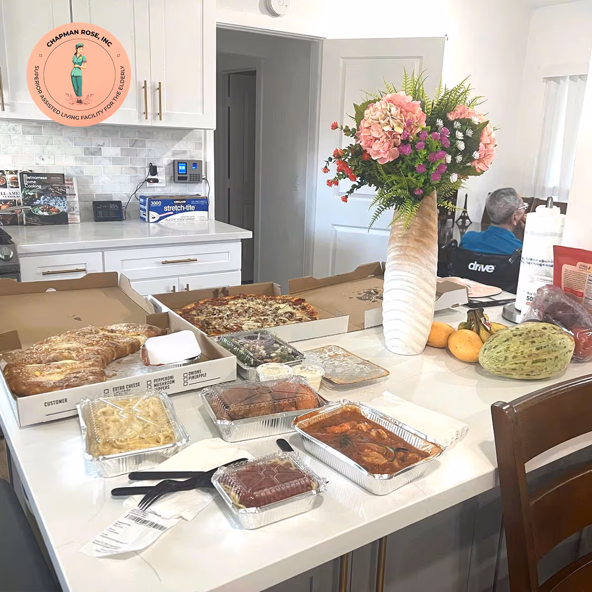 White kitchen counter with pizza boxes, assorted foods, and a tall floral arrangement.