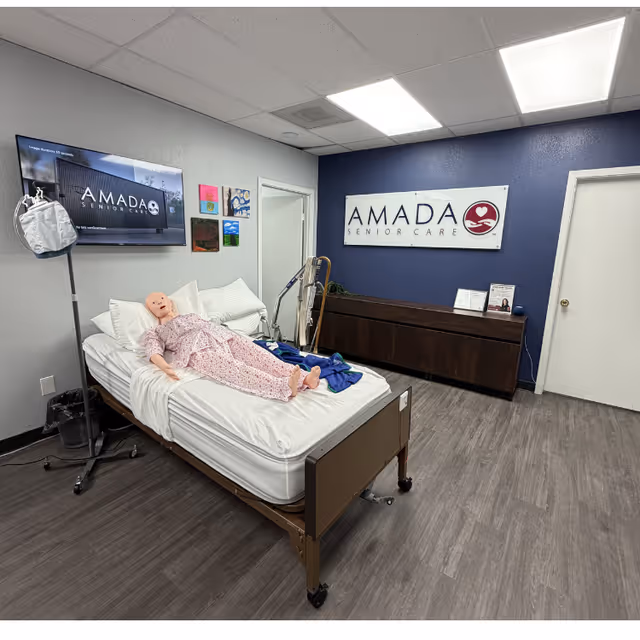 A patient bed with a mannequin lies in a facility room featuring Amada Senior Care signage.