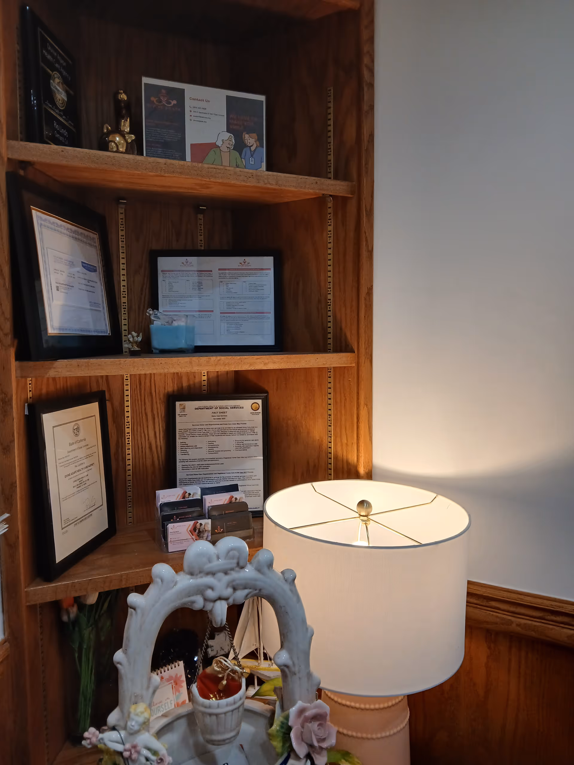 Wooden wall shelf with framed certificates, a decorative lamp, and trinkets in a lobby.