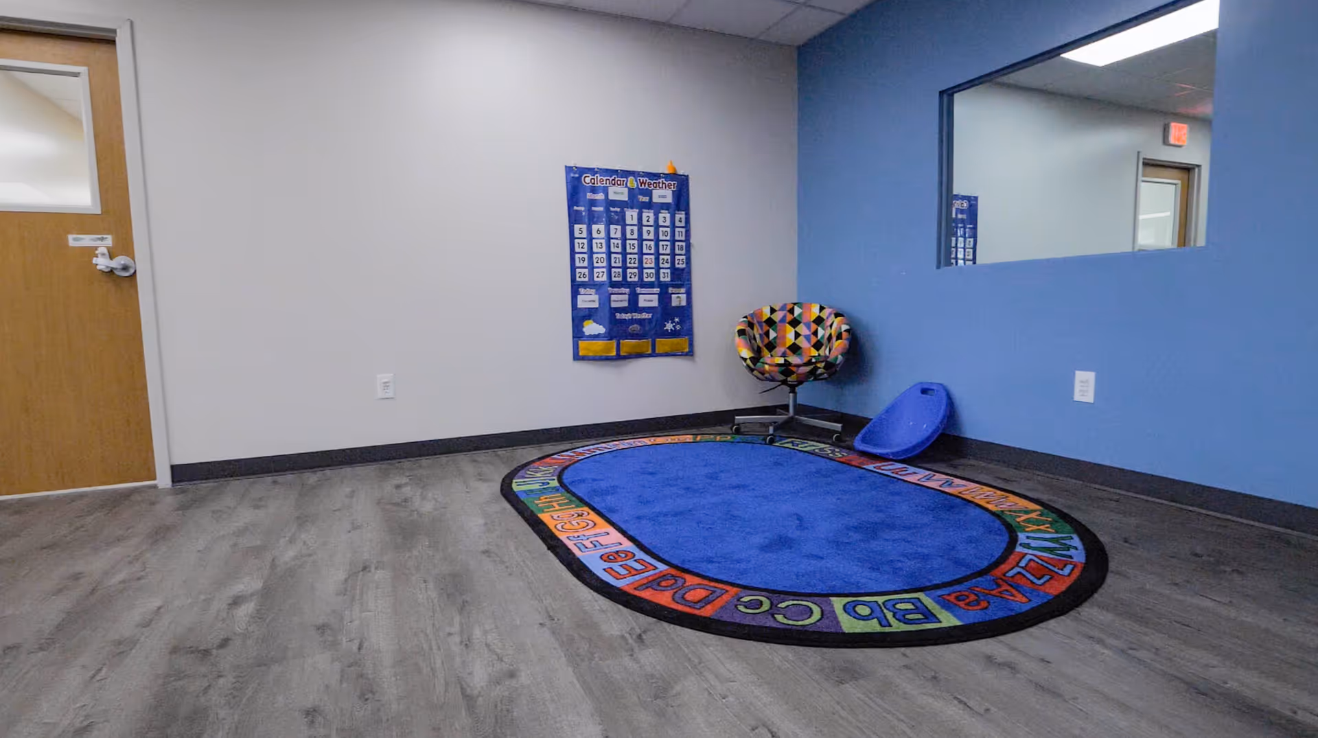 An empty indoor activity room with a blue wall, colorful oval rug, calendar, and patterned chair.