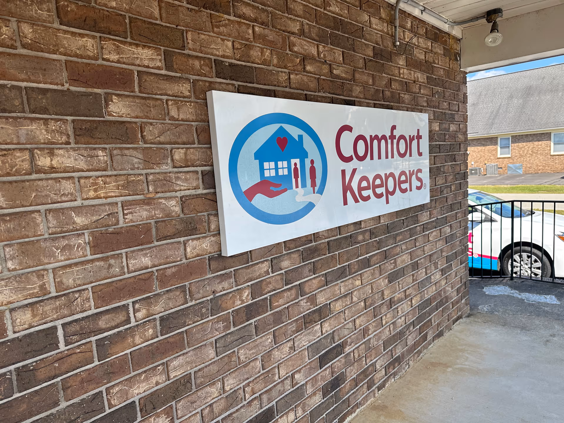 Sign reading 'Comfort Keepers' mounted on brick exterior wall by parking.